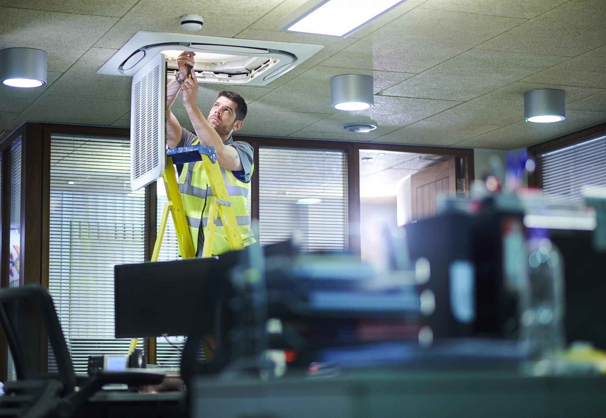 Male engineer on a ladder fixing an air conditioning unit in an office