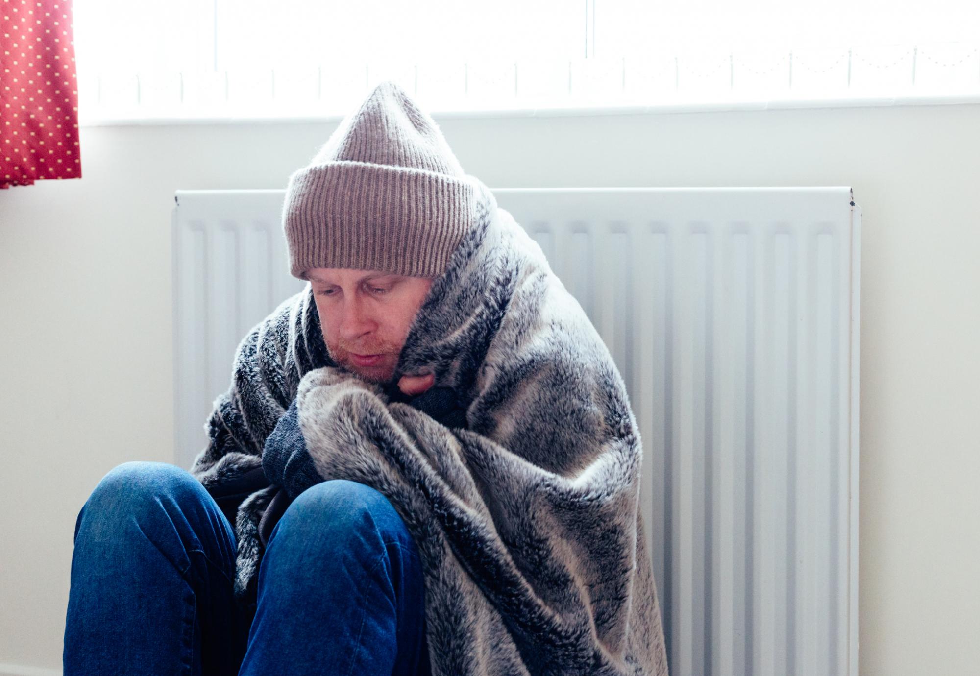 A cold mid adult man sits next to a warm radiator at home during winter
