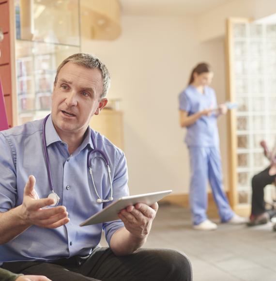Image of a health professional holding a digital tablet talking to a patient depicting the AI funding and IT infrastructure